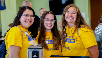 Three female students wearing Madonna University Peer Mentor shirts that are yellow smiling for the camera in the cafeteria 